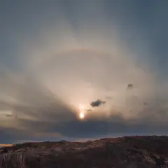 Słońce otoczone halo na tle chmur nad wzgórzem.