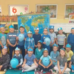 Grupa przedszkolaków w niebieskich czapeczkach z kropelkami wody, trzymających balony.