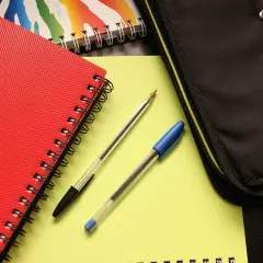 Czerwony zeszyt, dwa długopisy i piórnik na żółtym tle.