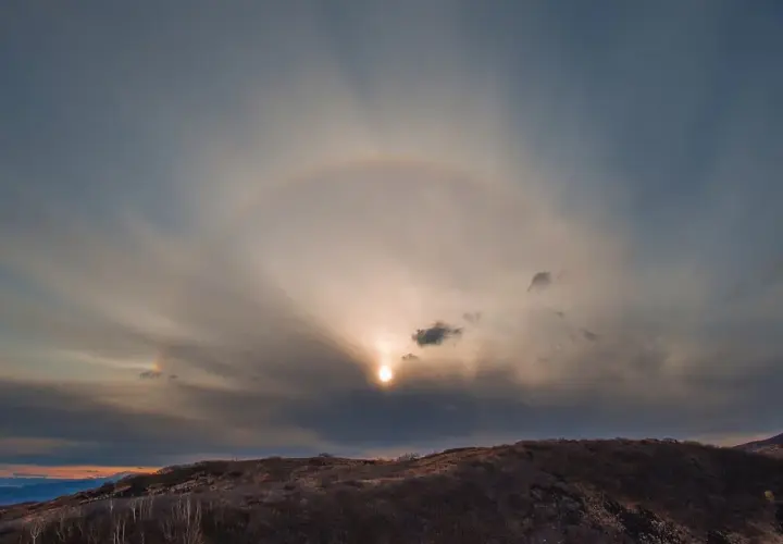 Słońce otoczone halo na tle chmur nad wzgórzem.