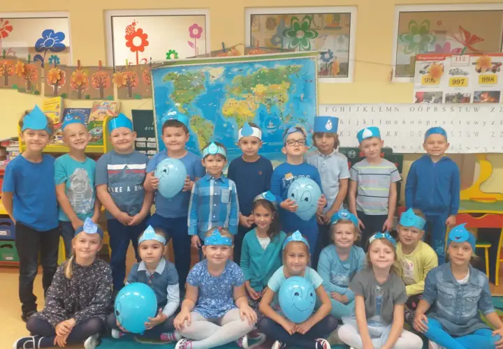 Grupa przedszkolaków w niebieskich czapeczkach z kropelkami wody, trzymających balony.