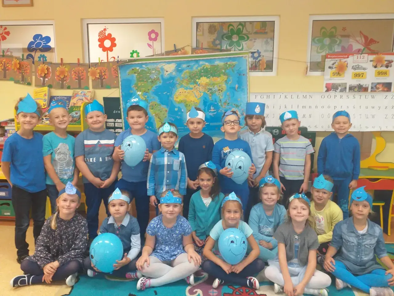 Grupa przedszkolaków w niebieskich czapeczkach z kropelkami wody, trzymających balony.